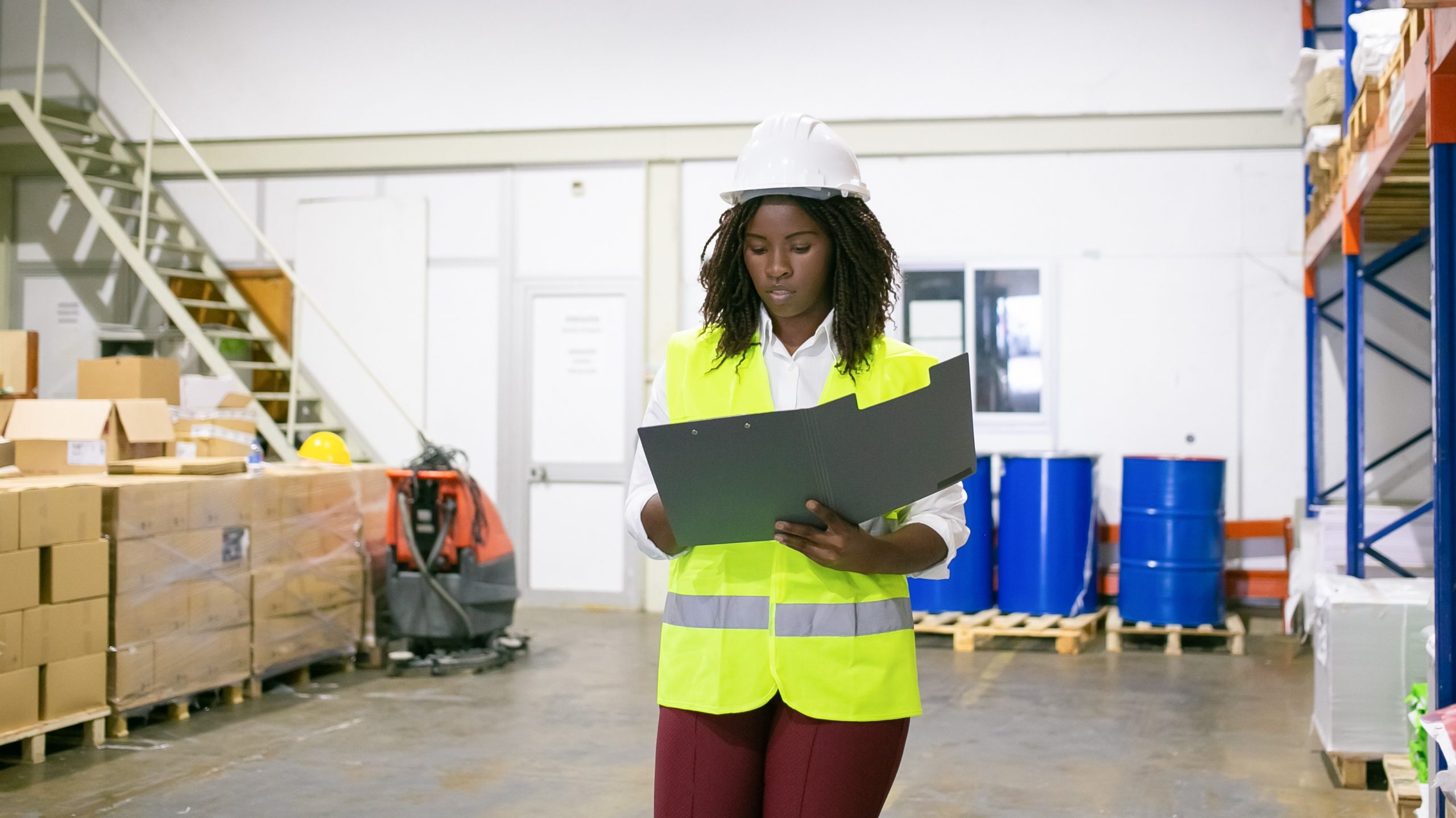 Focused female logistic employee in hardhat and safety vest walking in warehouse, carrying open folder, looking through document. Copy space, front view. Labor and inspection concept